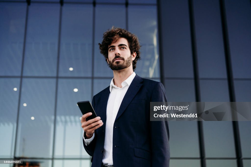 Contemplative businessman with smart phone in front of modern building