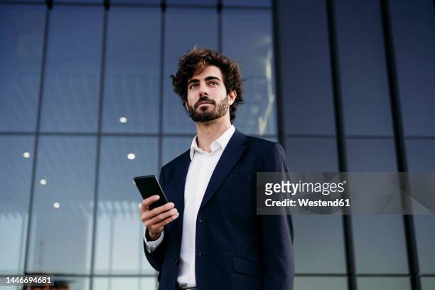contemplative businessman with smart phone in front of modern building - business man stock pictures, royalty-free photos & images