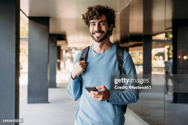 young handsome man with smart phone standing in corridor - ragazzo foto e immagini stock