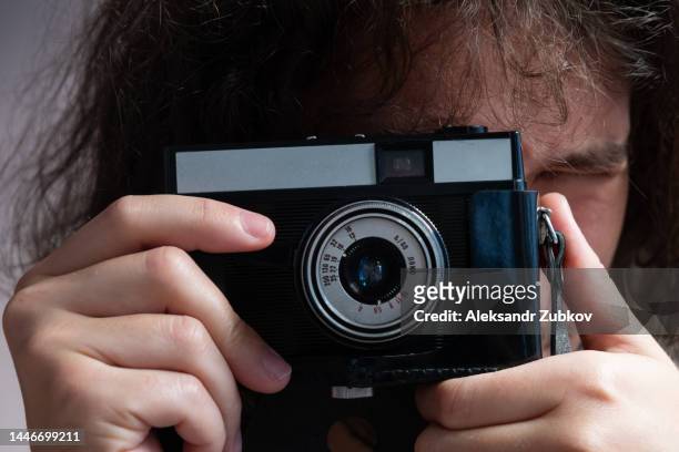 a man takes pictures with a retro camera, holds it in his hands. a young man, photographer, traveler or tourist working indoors. a creative freelancer, a student in casual clothes, takes a picture on camera. the concept of professional activity, hobby. - media day creative portraits stock pictures, royalty-free photos & images