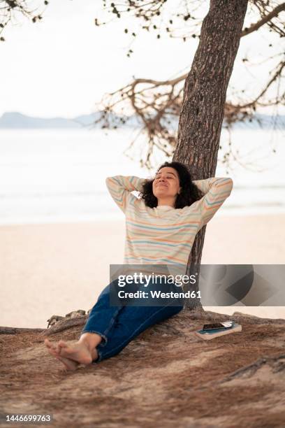 woman with hands behind head leaning on tree - füße überkreuzt stock-fotos und bilder