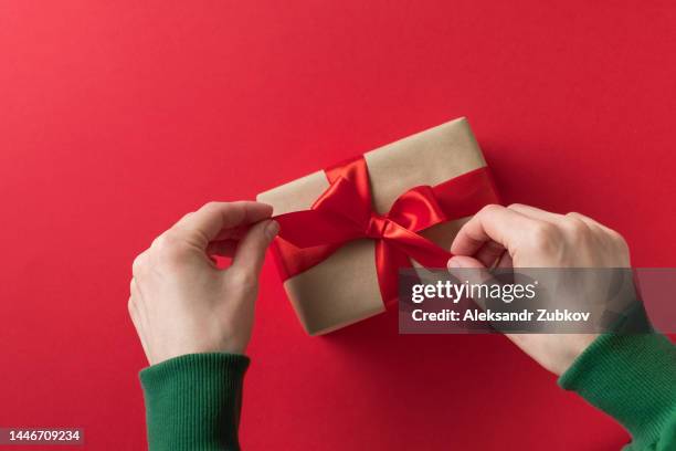 a girl in a green sweater holds a beautiful festive gift box with a surprise tied with a bright ribbon on a red background. a woman packs a gift or opens it on her holiday. the concept of merry christmas, birthday, valentine's day. copy space. - gift lounge stock-fotos und bilder