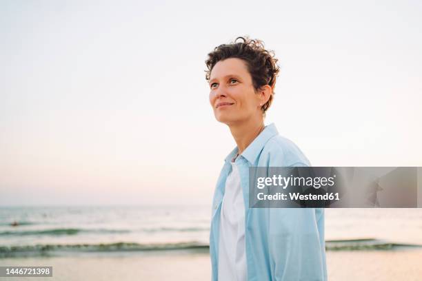 thoughtful androgynous woman standing at beach - short stock pictures, royalty-free photos & images