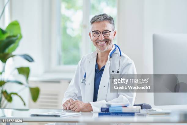 happy mature doctor in lab coat sitting in medical practice - doctor coat stock pictures, royalty-free photos & images