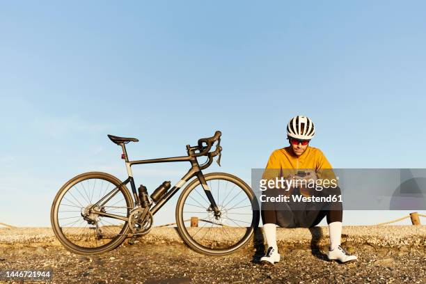 young cyclist using smart phone sitting under sky - sports helmet fotografías e imágenes de stock
