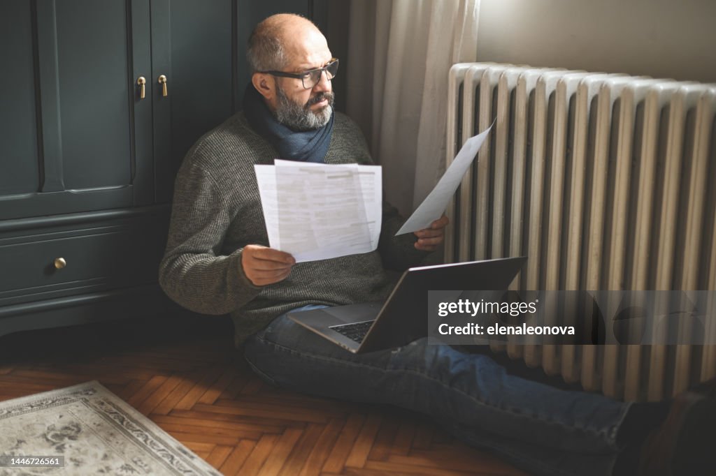 Reifer erwachsener Mann, der zu Hause arbeitet (Heizkörper, Laptop, Finanzrechnung)