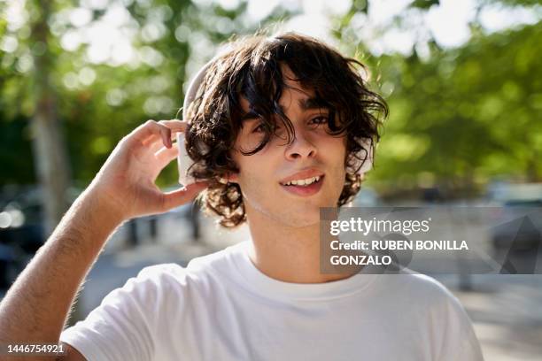 portrait of carefree smiling young caucasian teenager putting headphones on ears for listening to music outdoors in monumental street. music and lifestyle - boy headphones stock-fotos und bilder