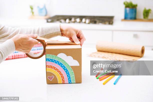 woman using brown tape to secure a handmade gift box with cute rainbow design. concept of shipping happiness. - care package stock pictures, royalty-free photos & images