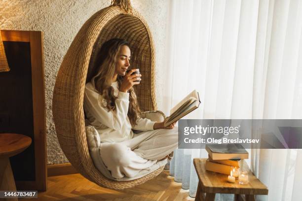 woman relaxing at home in hanging ball chair, reading book and drinking hot herbal tea - cocoon stockfoto's en -beelden