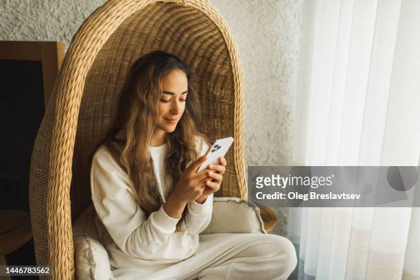young woman relaxing in hanging wicker chair with mobile phone. wasting time and procrastination - wicker - fotografias e filmes do acervo
