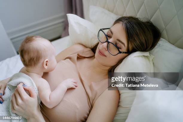 breastfeeding of little baby in bed. young mother in glasses embracing and feeding newborn baby in bed - best bosom stock-fotos und bilder