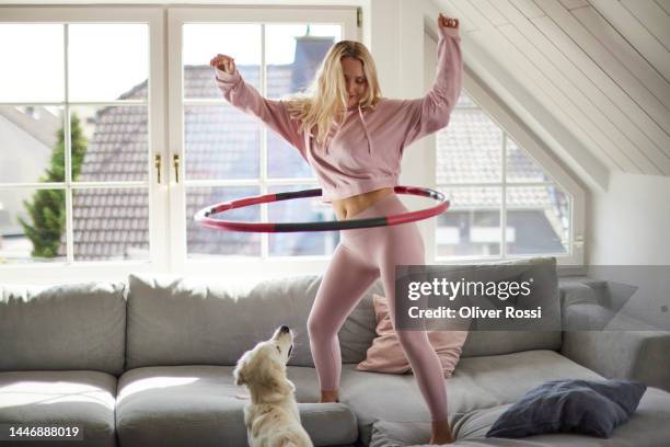 young woman using plastic hoop on sofa at home watched by her dog - gymnastikreifen stock-fotos und bilder