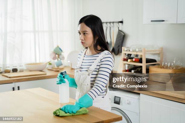 tired and mad asian woman spraying and cleaning the table with rag cloth. frustrated girl housekeeping and tidying up the kitchen - bored housewife stock pictures, royalty-free photos & images