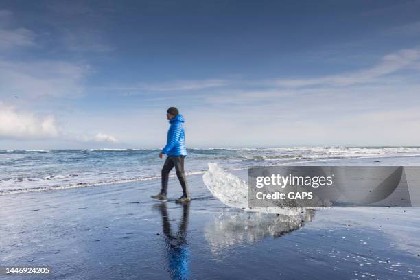 man in blue winter coat walking along the beautiful blue colored blocks of ice on the black sand of diamond beach in iceland - iceland people stock pictures, royalty-free photos & images