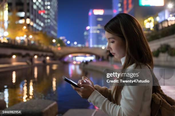 asian woman using a smartphone sitting at cheonggyecheon stream river park at night in seoul city - south korea people stock pictures, royalty-free photos & images