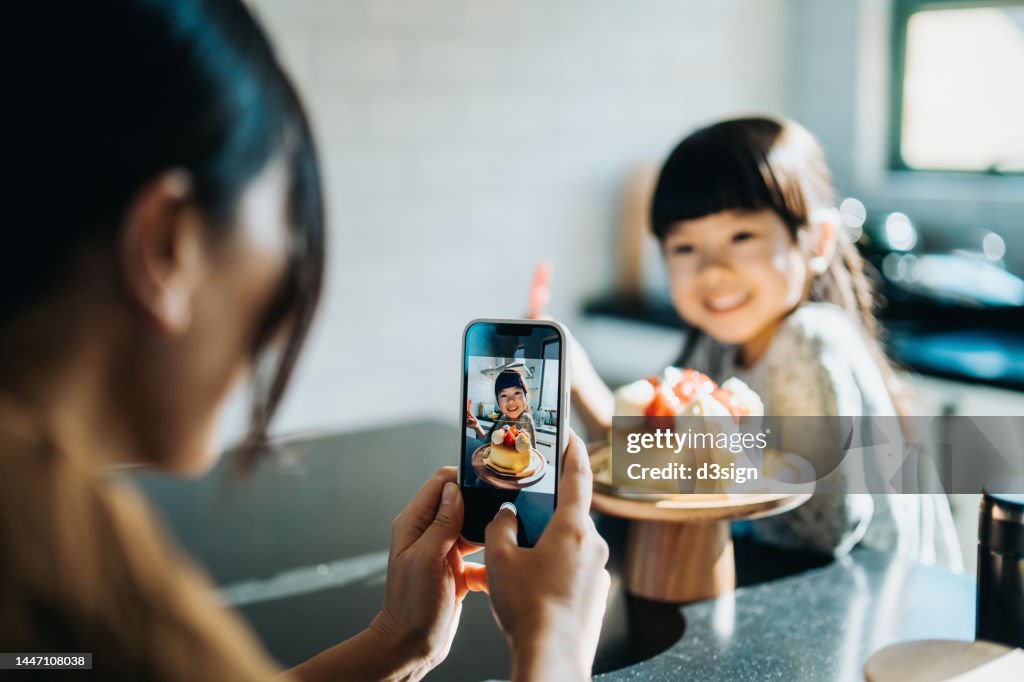 Cropped shot of young Asian mother celebrating 5th birthday with her lovely daughter, taking pictures with smartphone of the birthday girl holding a number 5 candle in front of the birthday cake. Birthday celebration and party theme