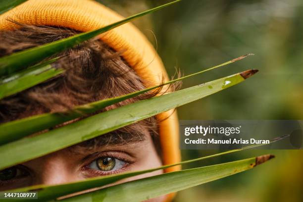 close-up of the eye of a teenager in a yellow cap behind a palm leaf - iris mann stock-fotos und bilder