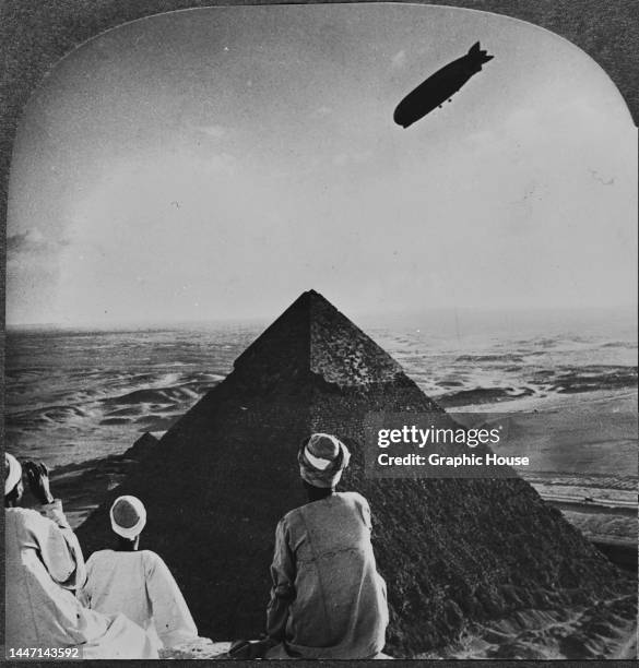 Rear view of Egyptian men in traditional clothing with a Graf Zeppelin in the sky over an pyramid, the Eternal Desert stretching out beyond, at Giza,...