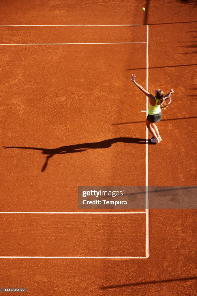 Vue aérienne d’une jeune femme adulte avec une ombre servant la balle de tennis sur un court en terre battue