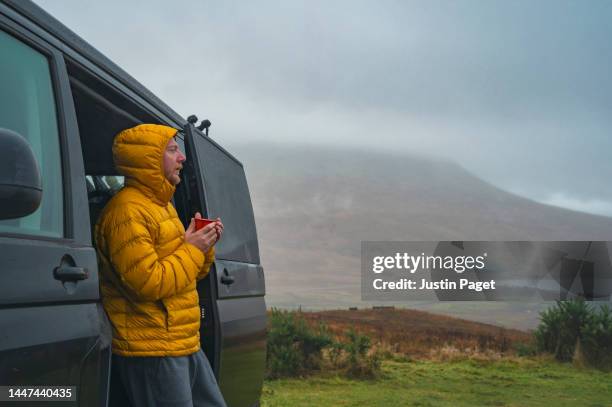 a mature man takes in the view of the scottish highlands on a rainy day from his campervan. he wears his hooded puffer coat and warms himself up with a cup of tea - motorhome winter stock-fotos und bilder