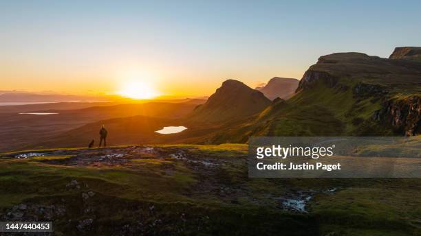 drone view of a man in red puffer coat taking in a beautiful sunrise view in the quiraing on the isle of skye. he's accompanied by his pet chocolate labrador dog - scotland people stock pictures, royalty-free photos & images