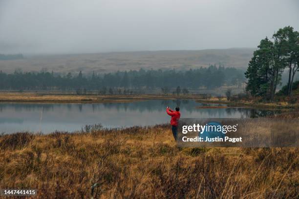 a mature man takes a photo of the beautiful misty view on his smartphone as he camps by a loch (lake) in scotland. - off the beaten path refrán en inglés fotografías e imágenes de stock