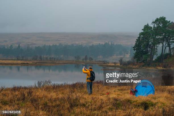 a mature man takes a photo of the beautiful misty view on his smartphone as he camps by a loch (lake) in scotland. - outdoor pursuit stock pictures, royalty-free photos & images