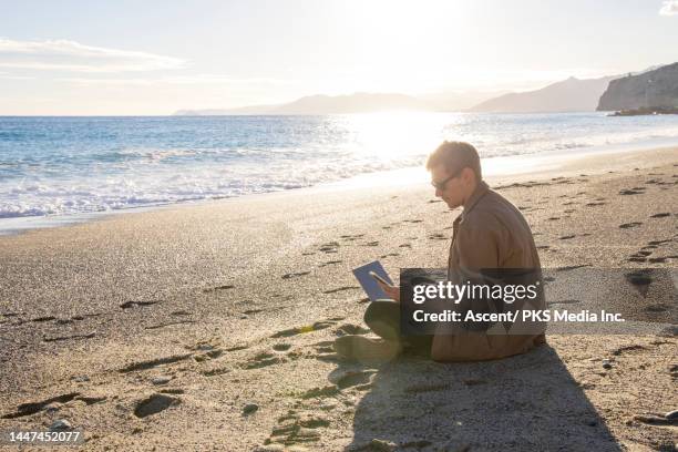 young man uses laptop on beach at sunset in autumn - moving image foto e immagini stock