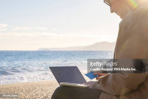 young man uses laptop makes digital payment on beach at sunrise - profileren stockfoto's en -beelden