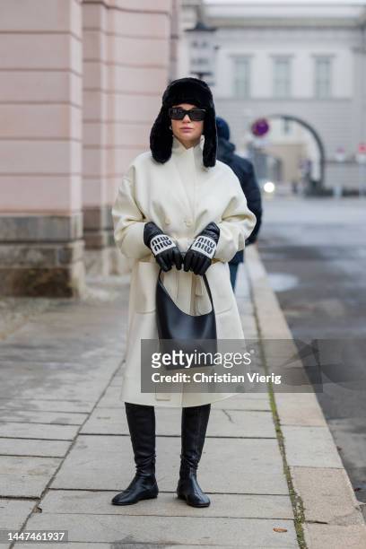 Alice Fetisova wears black heat, creme white belted coat, Miu Miu gloves, black bag, boots, sunglasses on December 06, 2022 in Berlin, Germany.