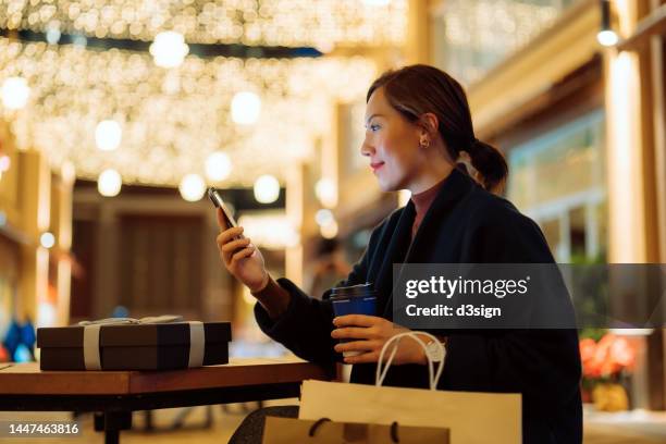 beautiful young asian woman relaxing on a bench and checking smartphone after shopping in the city, with decorated high street and illuminated christmas lights in background. enjoying christmas shopping. festive vibes. christmas is almost here - woman shopping china photos et images de collection