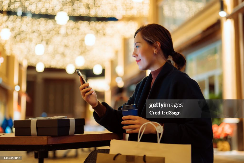 Beautiful young Asian woman relaxing on a bench and checking smartphone after shopping in the city, with decorated high street and illuminated Christmas lights in background. Enjoying Christmas shopping. Festive vibes. Christmas is almost here
