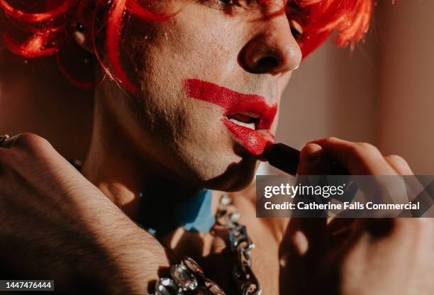 a man wearing a wig applies lipstick, intentionally smudging it around his lips. - beautiful transvestite stock pictures, royalty-free photos & images