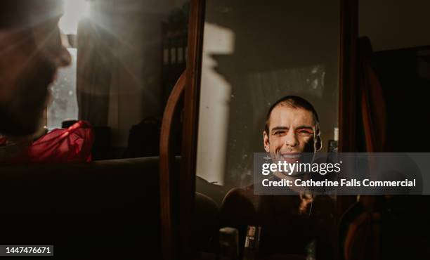 a man wearing orange eye make-up and red lipstick looks in a mirror and smiles at himself. he is confident. - beautiful transvestite stock pictures, royalty-free photos & images