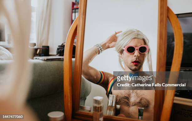 a man wearing sunglasses and a blond wig admires his look in a vanity mirror - applying make up stockfoto's en -beelden