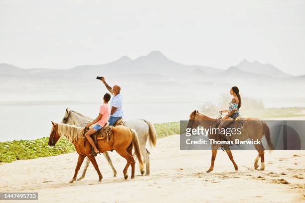 Wide shot of family taking selfie while riding horses on tropical beach