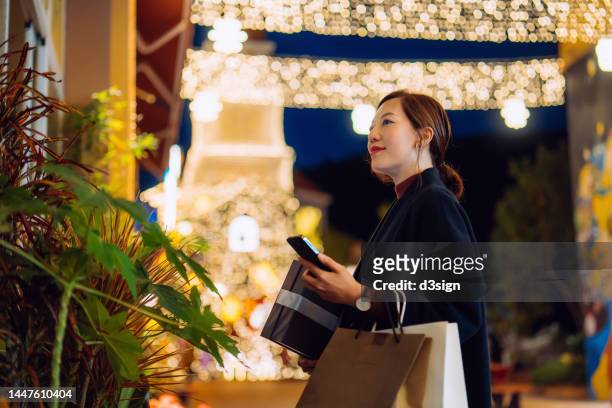 young asian woman with smartphone window shopping in decorated city street with illuminated christmas lights. looking at window display through store window. holding christmas presents. enjoying christmas shopping. festive vibes. christmas is almost here - angeleuchtet zahlen mensch stock-fotos und bilder