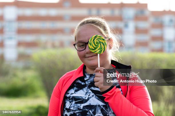 portrait of a chubby blonde girl covering her eye with a lollipop. - childhood obesity stock pictures, royalty-free photos & images