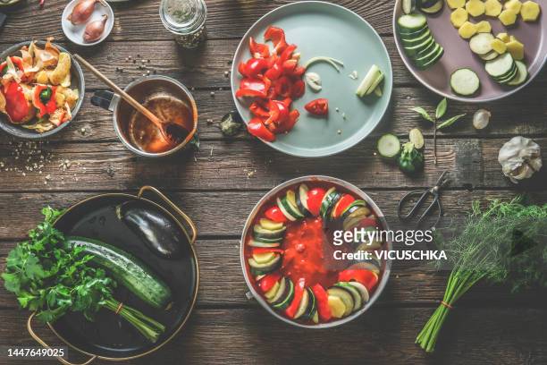 coking preparation of vegan ratatouille with vegetables ingredients, cooking pan, herbs, spices, tomato sauce and kitchen utensils on dark wooden background - ratatouille stock pictures, royalty-free photos & images