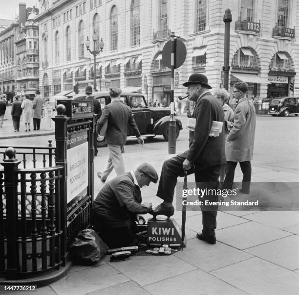 Man getting his shoes shined at Piccadilly Circus in London on May 21st, 1962. The advertising sign on the shoeshine man's box reads 'Kiwi Polishes'.