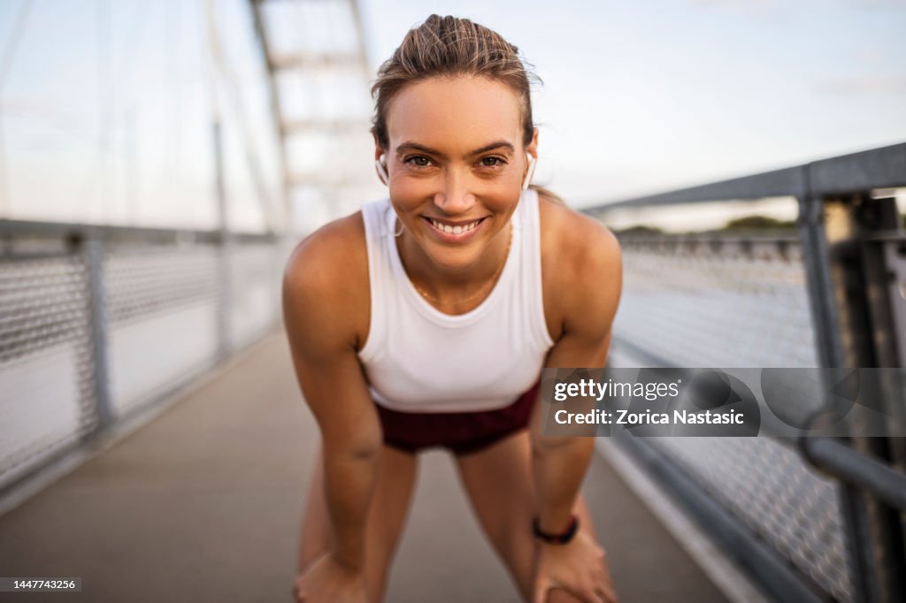 Young sportswoman taking a break after running on the bridge looking at camera smiling