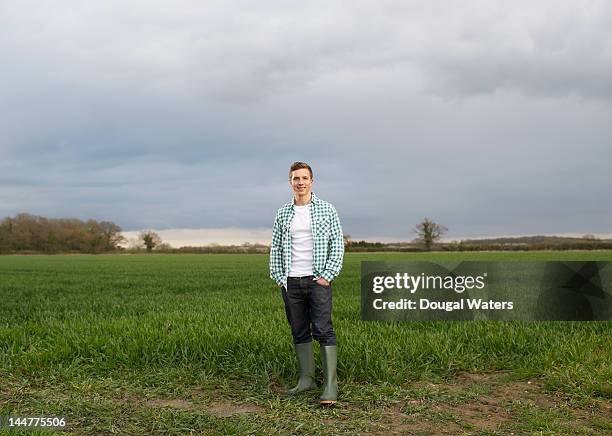 young farmer standing at edge of field. - bottes photos et images de collection