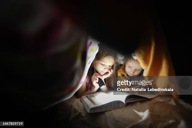 childhood moments. two sisters hide under blanket and read book with flashlight. dark style and copy space. - kinderbuch stock-fotos und bilder