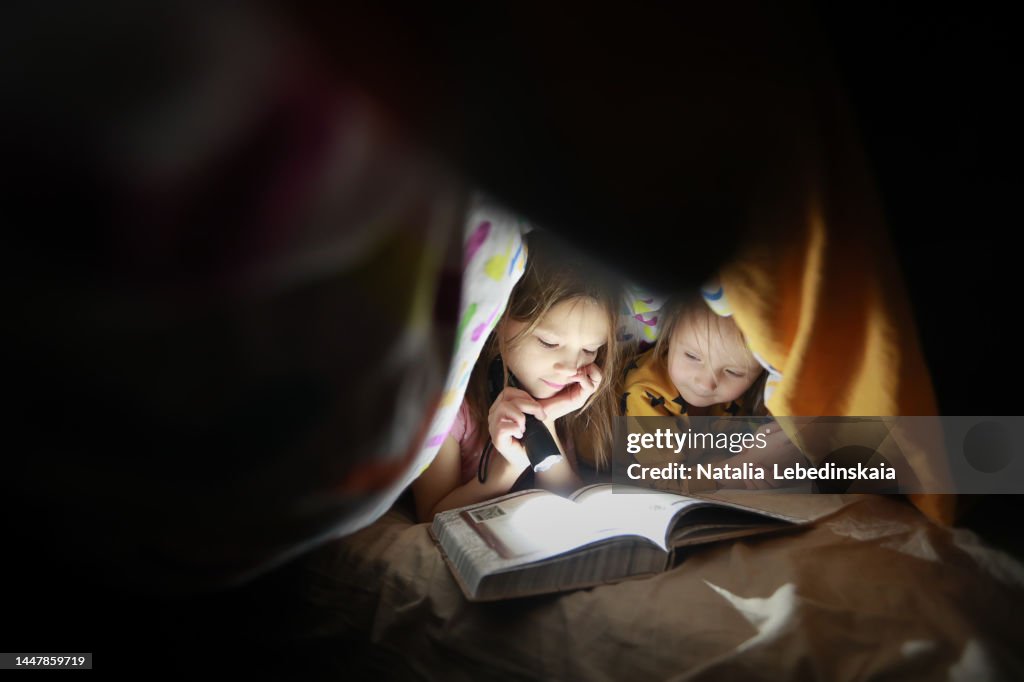 Childhood moments. Two sisters hide under blanket and read book with flashlight. Dark style and copy space.