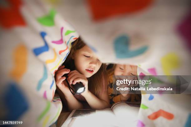 childhood moments. two sisters hide under blanket and read book with flashlight. close up. - manta-ropa-de-cama fotografías e imágenes de stock
