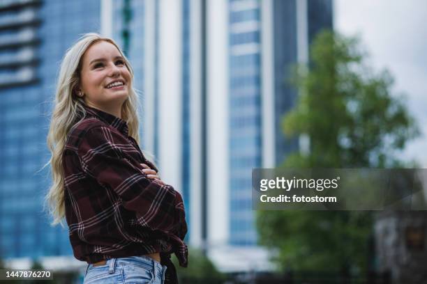 mujer joven de pie con los brazos cruzados, mirando hacia arriba, sonriendo y reflexionando - blond hair girl fotografías e imágenes de stock