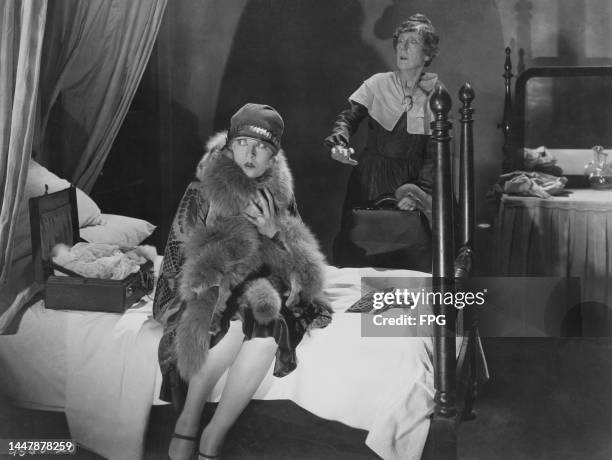 American actress Laura La Plante sitting on a bed beside an open suitcase, with American actress Martha Mattox in the background in a publicity still...