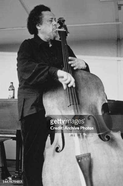 American jazz musician and composer Charles Mingus , a cigar in his mouth, playing a double bass, a piano partially visible behind him, performing on...