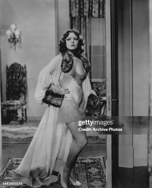 American actress Clara Bow wearing a fur-trimmed robe over a lace-trimmed slip, her hand on her hip, standing in white high-heel shoes in a bedroom,...