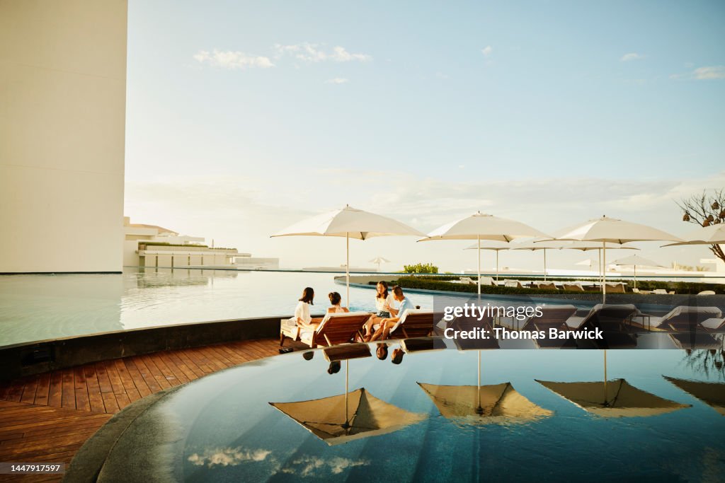 Wide shot of family hanging out on lounge chairs by pool at resort